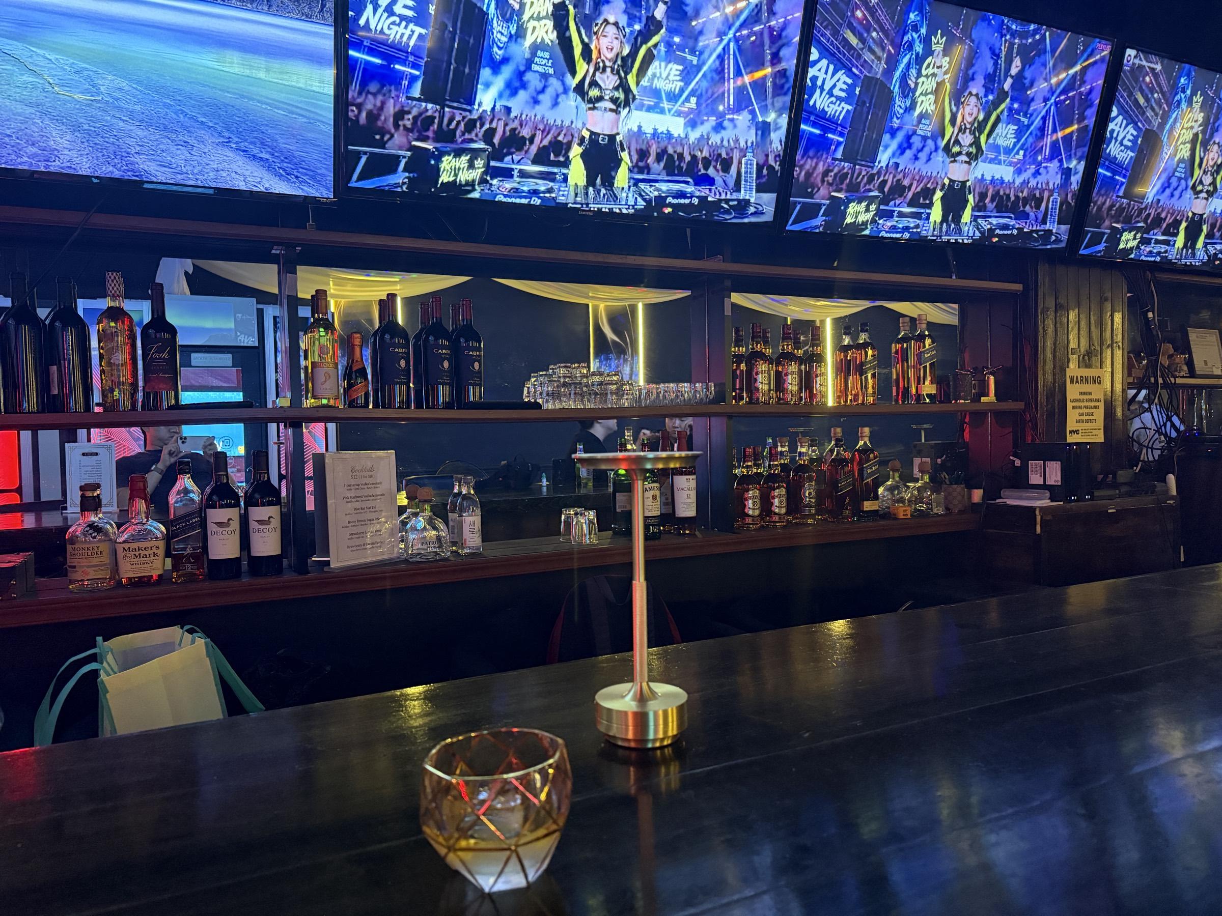 The bar at Bar Vier: bottles lit along the back shelves beneath a row of screens, with a gold-rimmed glass and a brass lamp on the dark wood counter.